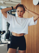 Kobieta w białym crop topie After Class i czarnych szortach na tle drewnianej ściany